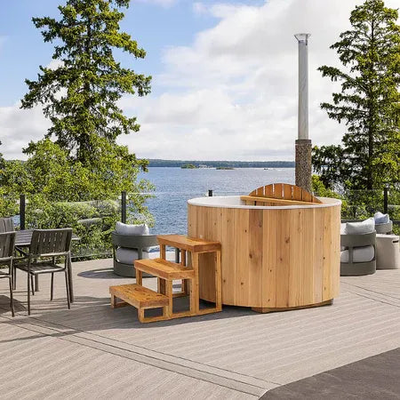 Handcrafted wooden hot tub by the water’s edge, featuring natural wood exterior and white interior for a serene lakeside retreat
